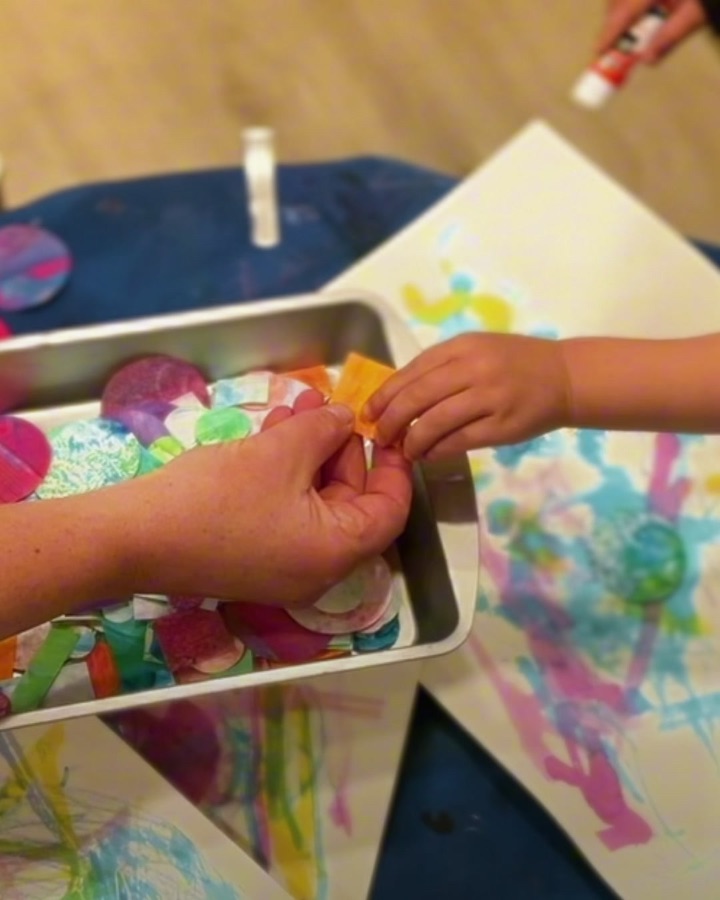 A teacher's hand and a child's hand reaching into a tray of colorful collage materials above paint-covered artwork, in a real Brooklyn preschool classroom